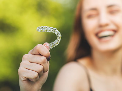 The image shows a person holding up a toothbrush with a smile, against a blurred outdoor background.