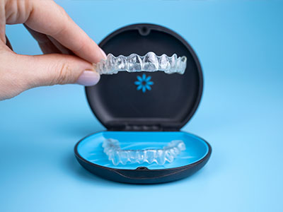 The image shows a hand holding an open clear case containing transparent dental braces with visible elastic bands, placed against a blue background.