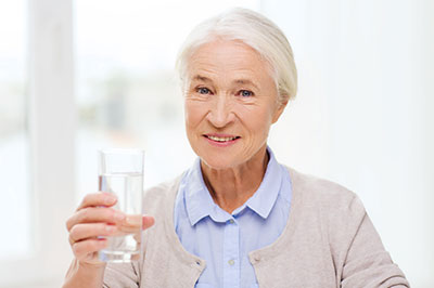 The image shows an elderly woman holding up a glass of water with both hands, smiling at the camera.