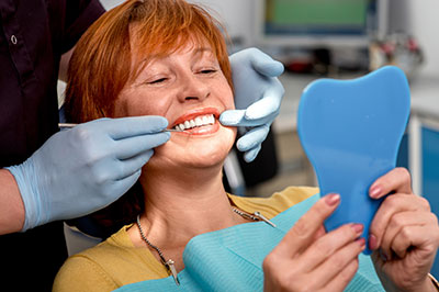 A woman with red hair is sitting in a dental chair, smiling at the camera while holding a blue dental model, with a dentist adjusting her teeth using a mirror and tools.