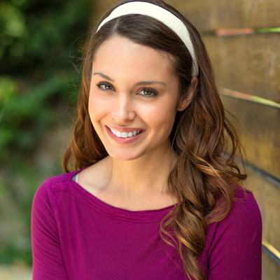 A smiling woman with long hair wearing a purple top and a headband poses against a wooden fence.