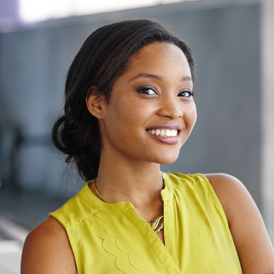 A smiling woman with dark hair, wearing a yellow top, poses against a gray background.