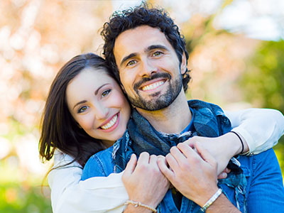 The image shows a man and woman embracing each other with smiles, indicating a close relationship, possibly romantic, set against an outdoor background.