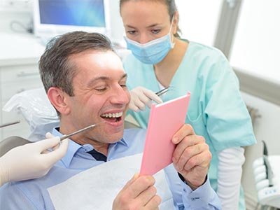 A man with a wide smile is holding up a pink card, seemingly showing it to others, while seated in a dental chair surrounded by dental professionals in a clinical setting.