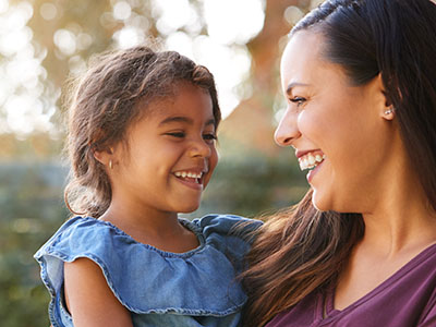 The image shows a woman and a young child smiling at each other outdoors during daylight.