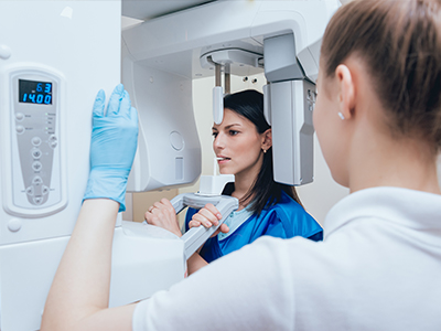 A woman in blue scrubs is standing next to a large 3D imaging machine while another person looks at her from behind, both are in a room with medical equipment.