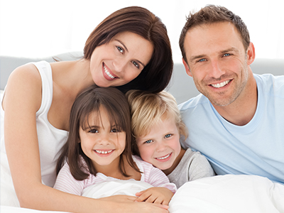 The image shows a family of four with two adults and two children, posing together for the camera with smiles on their faces, set against a plain background.