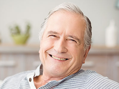 The image depicts an older man with gray hair, wearing glasses, smiling broadly, and leaning back slightly in a relaxed pose. He appears to be sitting in a home environment, possibly a living room, indicated by the presence of a bookshelf and other domestic items.