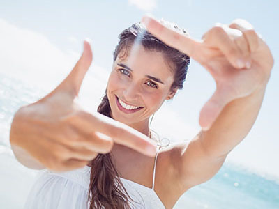 A woman with blonde hair and blue eyes is smiling at the camera while holding up her index finger against a bright, sunny background.